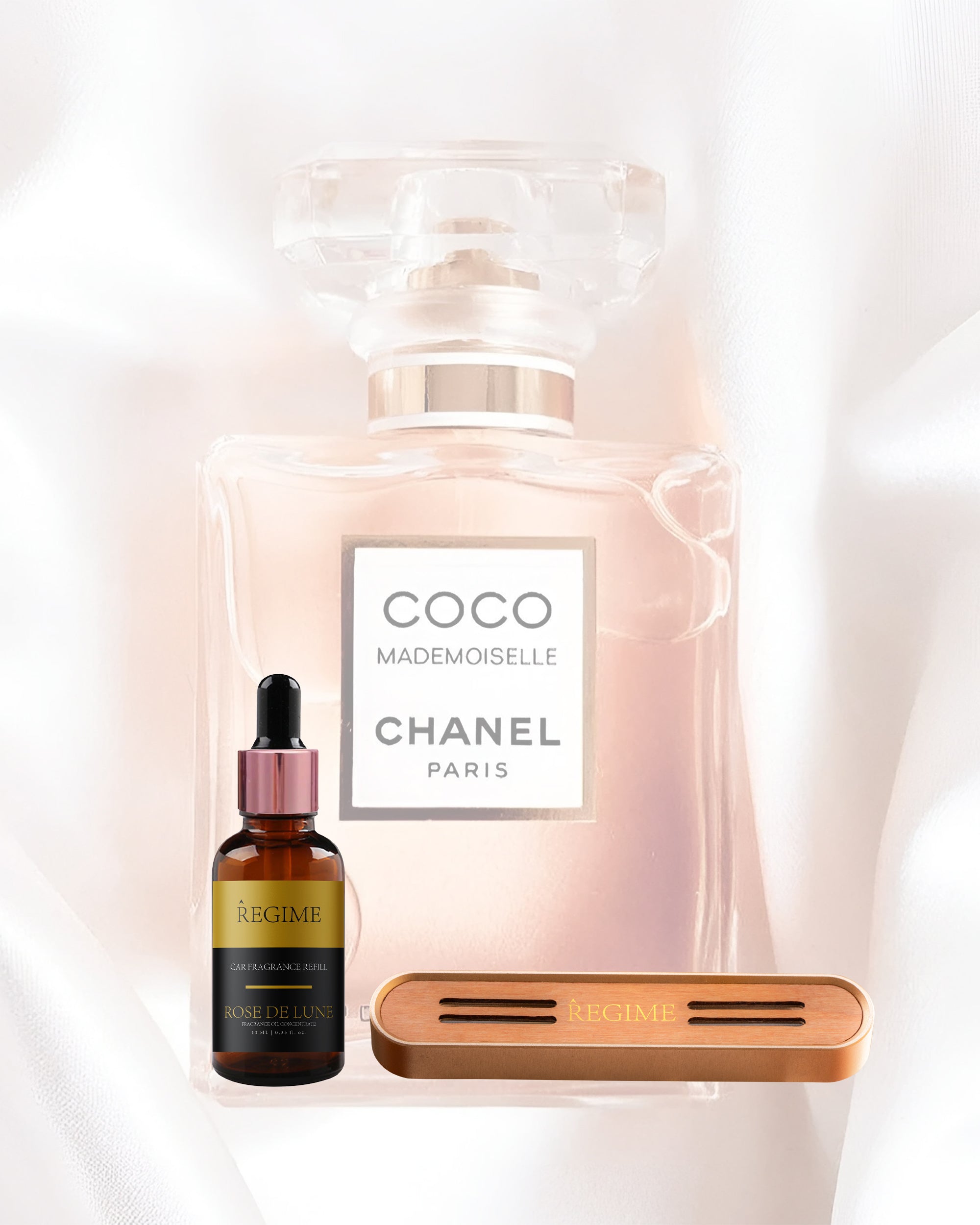 A faint image of Coco Chanel Mademoiselle perfume for women in the background; in the foreground, a fragrance oil dropper bottle inspired by Coco Chanel Mademoiselle and a rose gold metal Regime Fragrances car vent clip with a slotted wooden front panel for scent release.
